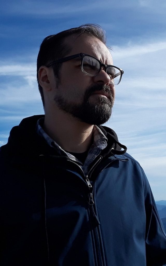 A man with dark hair, beard and glasses standing in front of a cloudy sky.